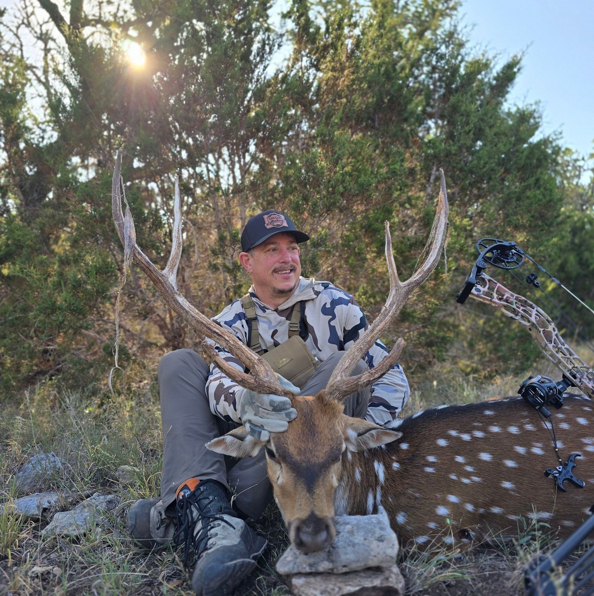 Hunter posing with a deer in a wooded area, wearing camouflage and a cap.