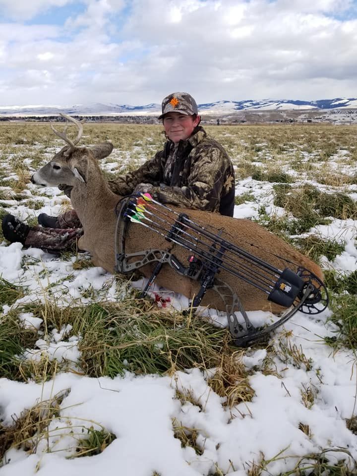 Hunter kneeling beside a deer in a snowy field, with a bow and gear visible.