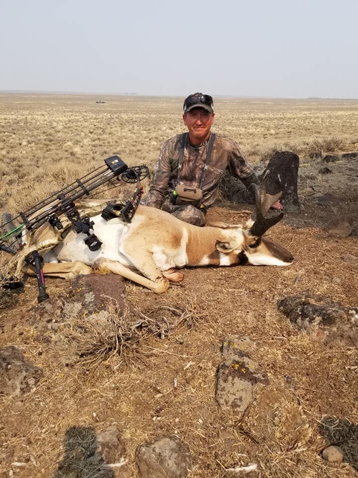 Hunter kneeling beside two hunted animals and a rifle in a dry open field