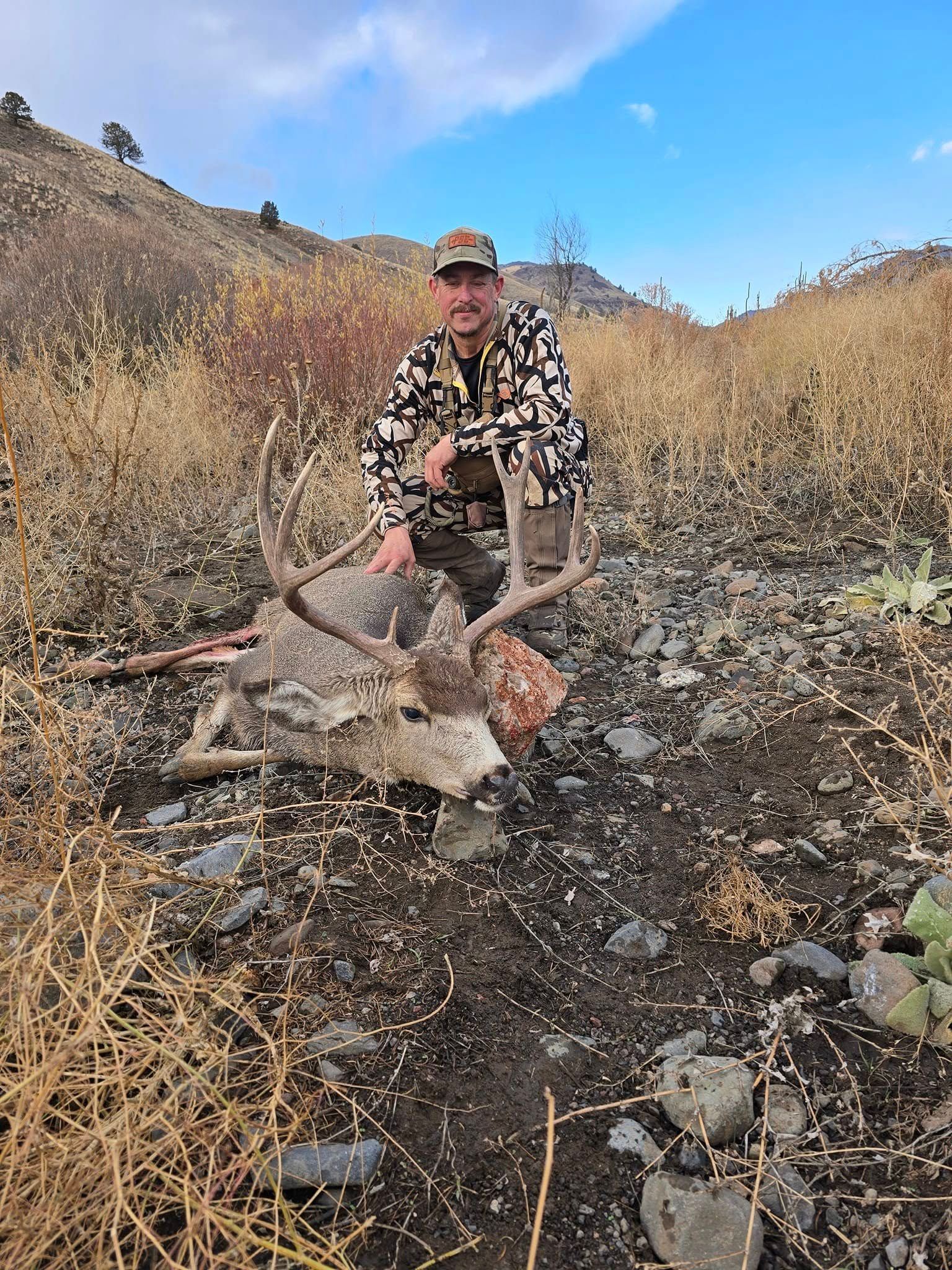 Person in camouflage crouches beside a dead deer in a dry, rocky hillside landscape.