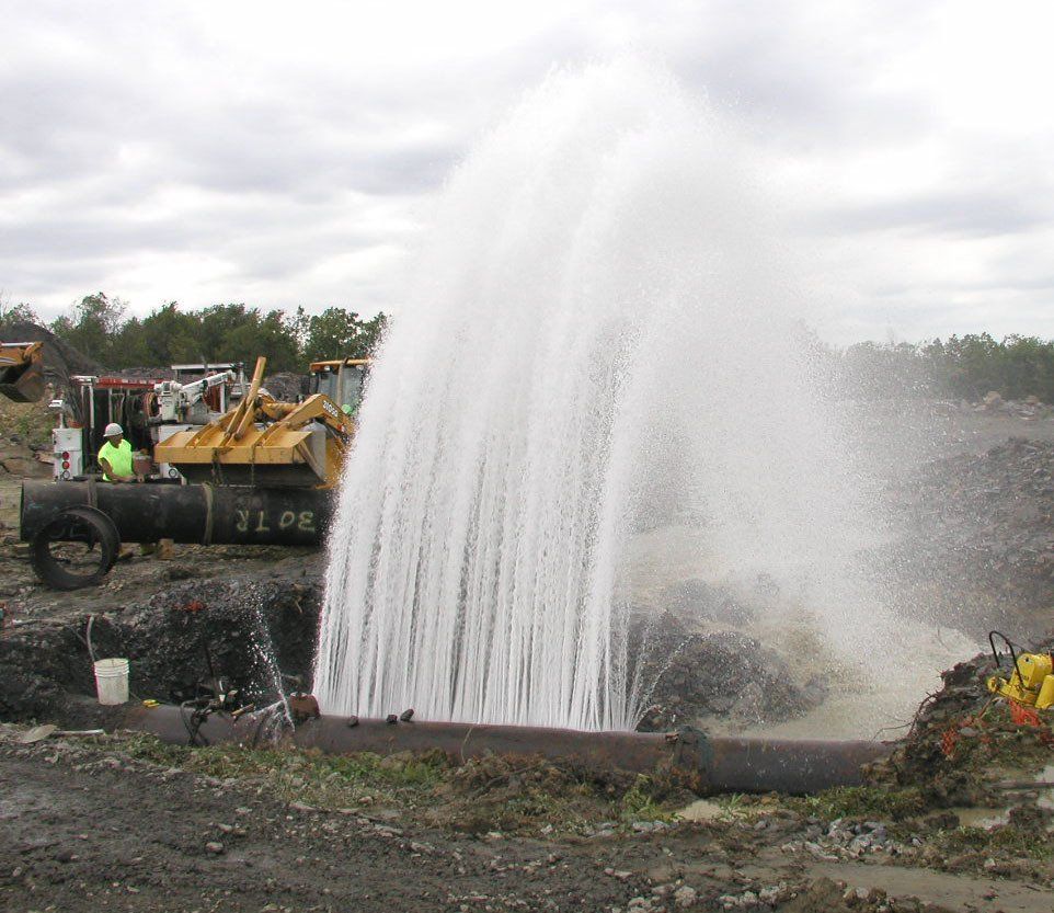 Water sprays upward from an underground pipe at a construction site.