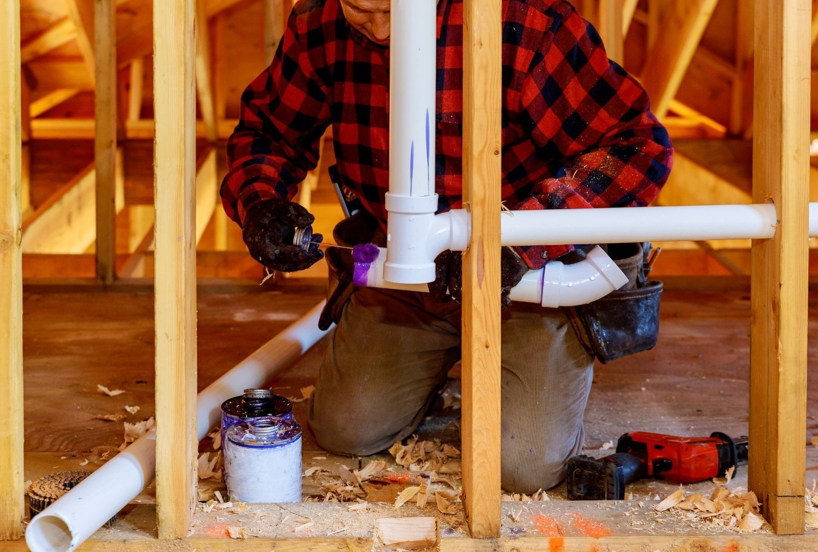 A man in a plaid shirt is working on pipes in a house under construction.