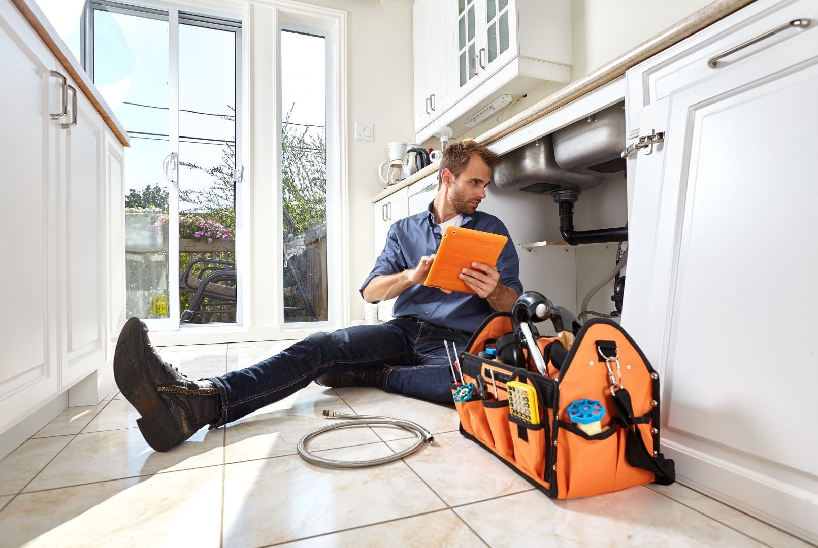 A plumber is sitting on the floor in a kitchen under a sink looking at a clipboard.