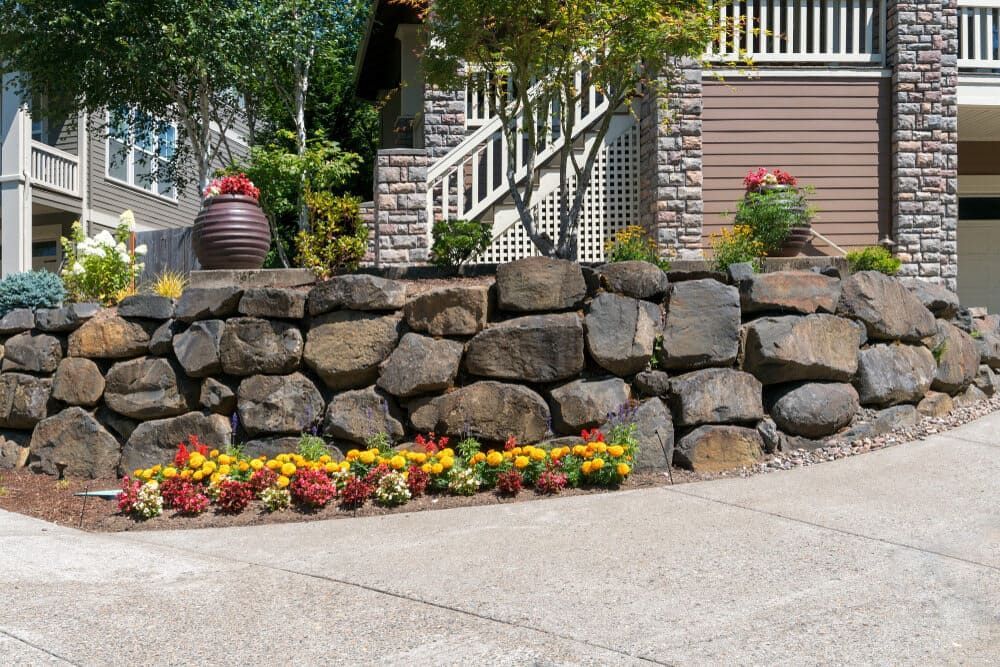 A Stone Wall With Flowers In Front Of A House — Baker & Co. Landscape Construction In Gulliver, QLD