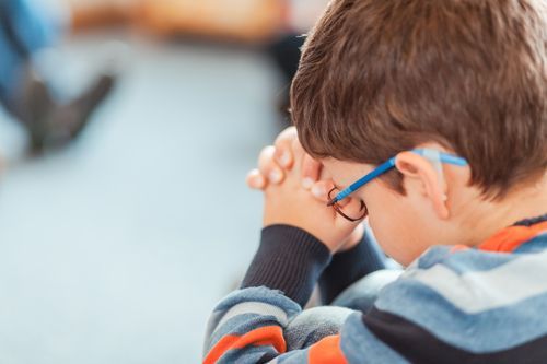 A young boy wearing glasses is praying with his hands folded in front of his face.