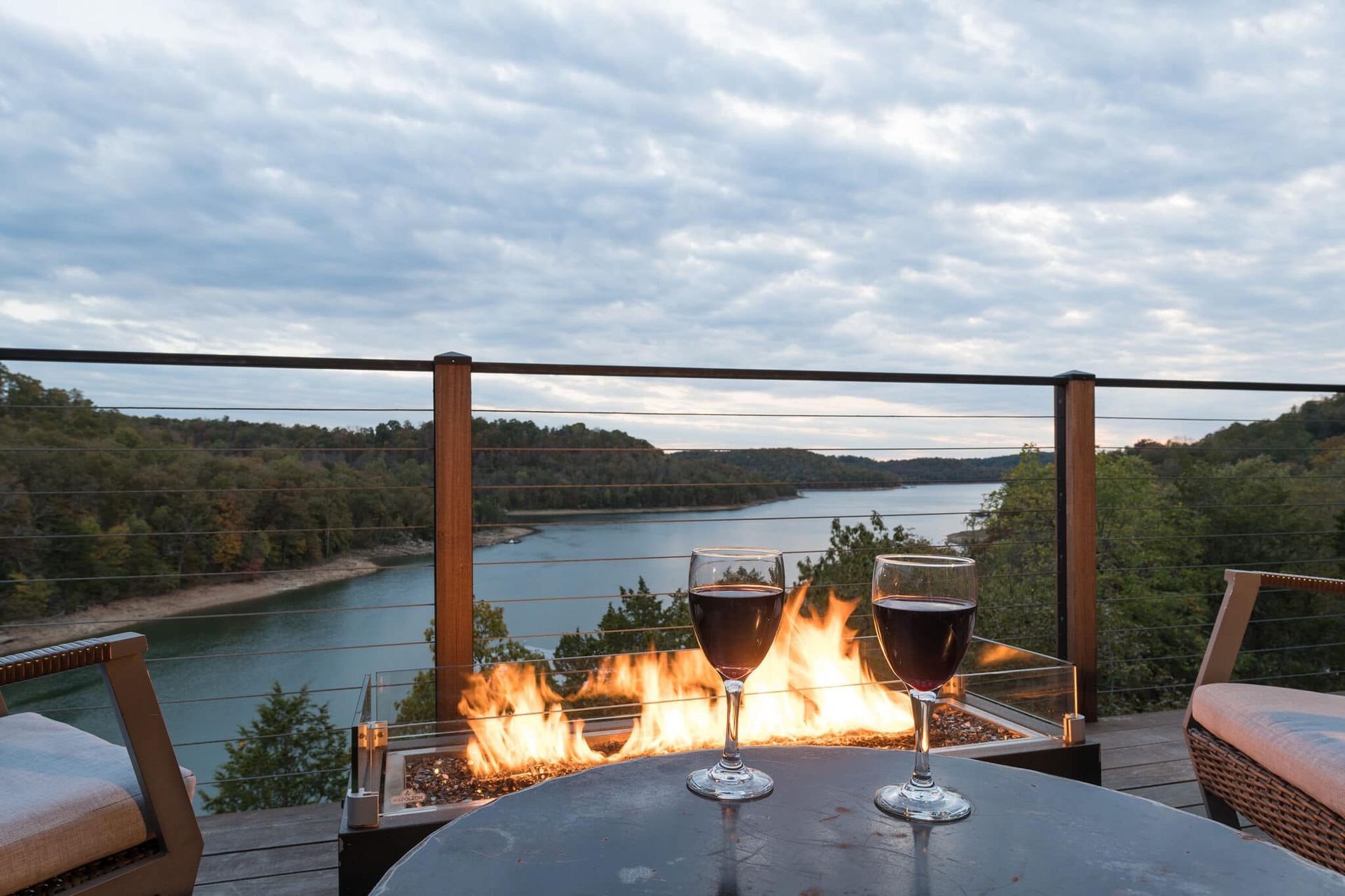 Two glasses of wine are sitting on a table next to a fire pit overlooking a lake.