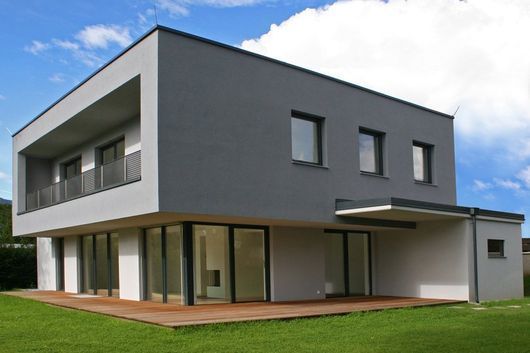 Casa moderna grigia a due piani con ampie finestre, balcone e prato verde.