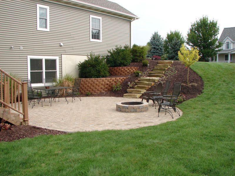 Backyard patio with fire pit, retaining walls, steps, and landscaping near a house with green lawn.