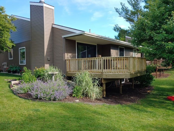 A brown house with a wooden deck, lush garden, and green lawn under a clear blue sky.