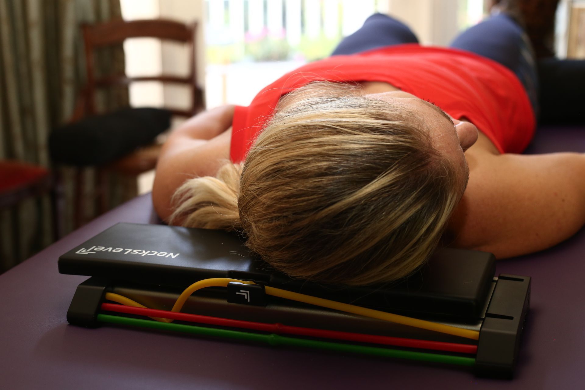 Woman lying on a massage table with head resting on a neck support device. Red shirt, blond hair.