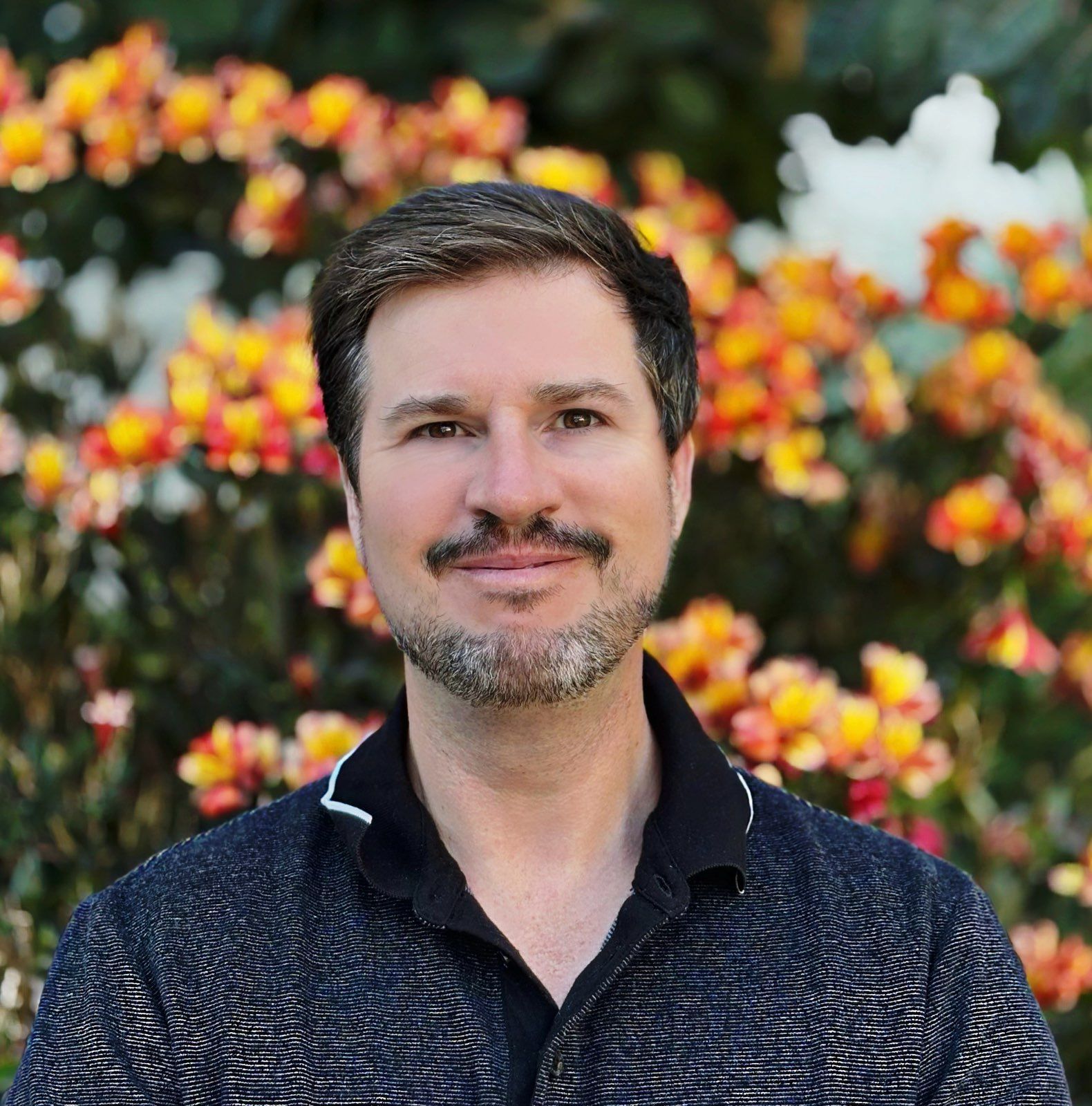 Man with a light beard smiles in front of colorful orange and yellow flowers.