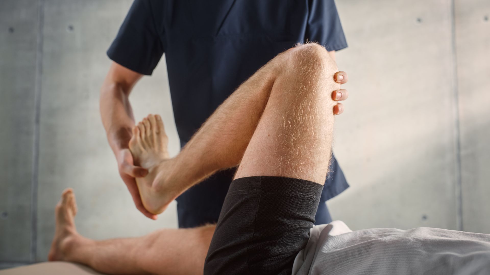 Physiotherapist manipulating a patient's leg, assessing knee movement. Indoor setting, gray background.