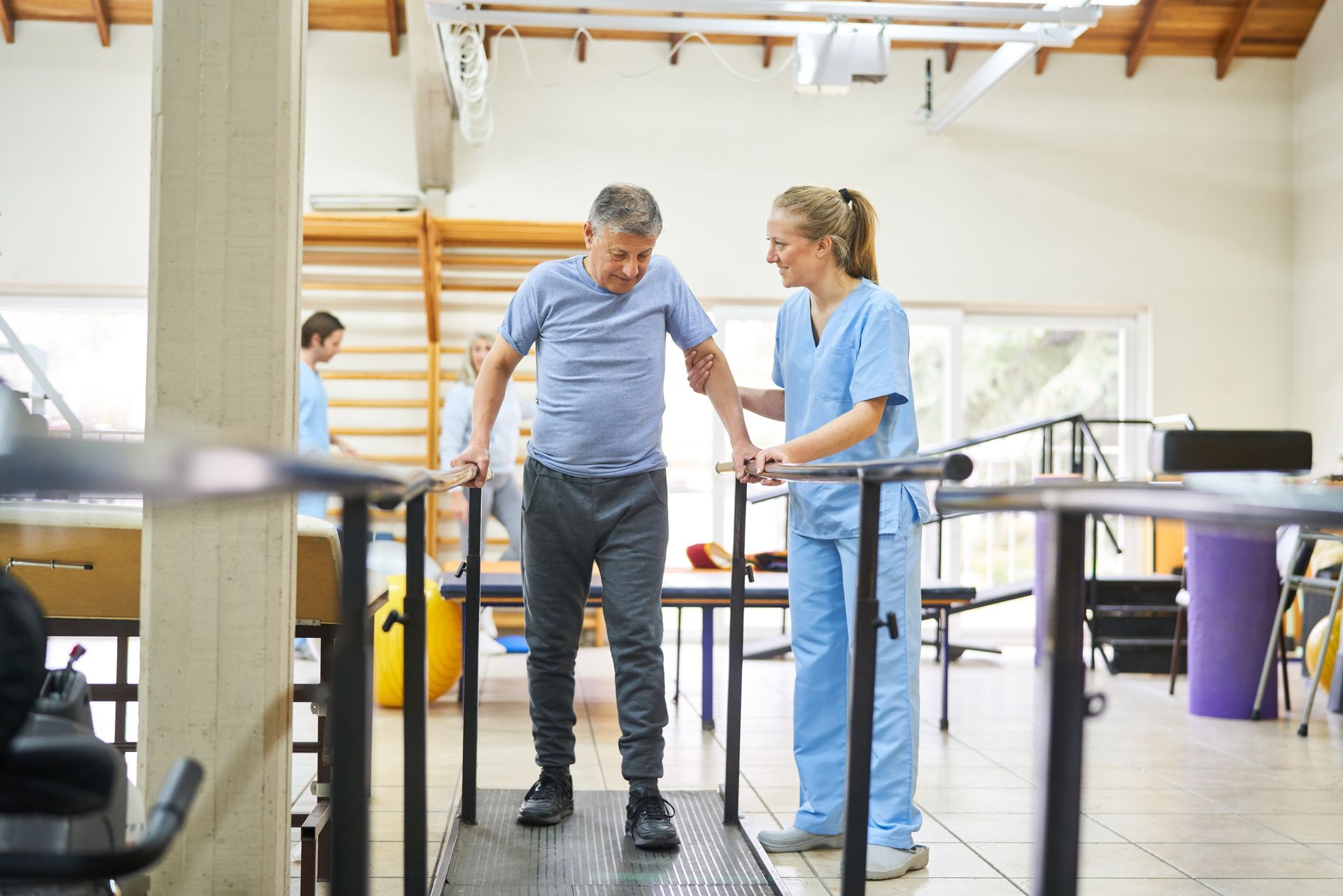 Man in rehab walking on treadmill, assisted by a female therapist in a light-filled room.