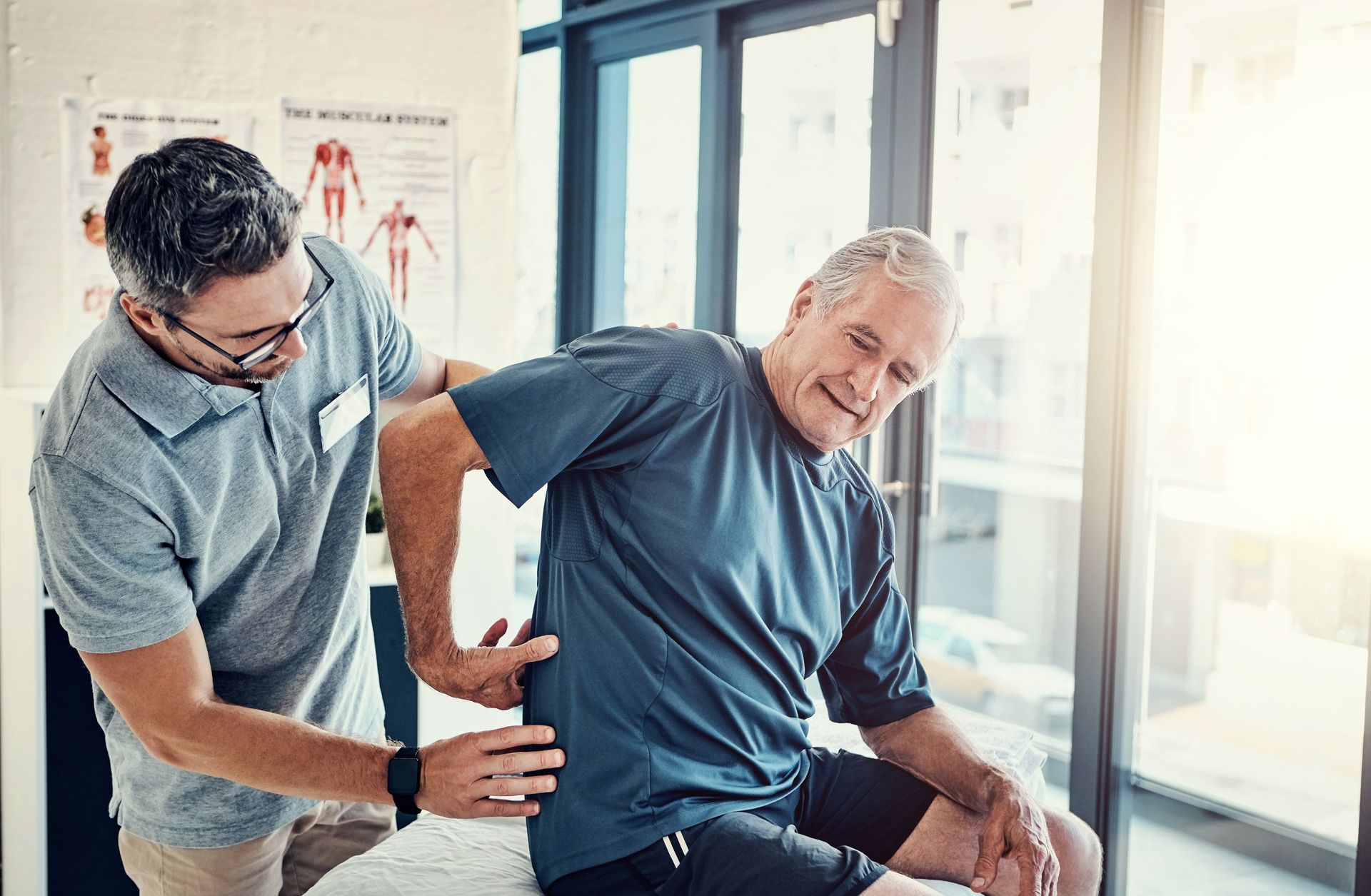 Physical therapist assisting an elderly man with back pain, indoors near a window.