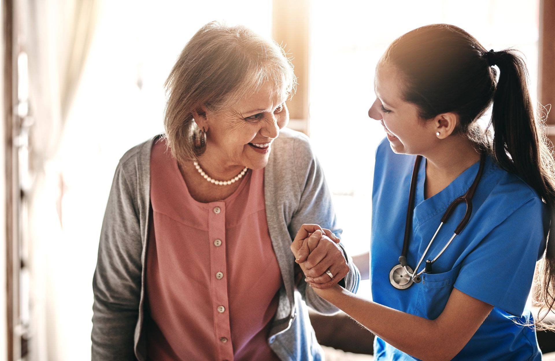 A smiling nurse helps an elderly woman walk, holding her arm, indoors in bright sunlight.