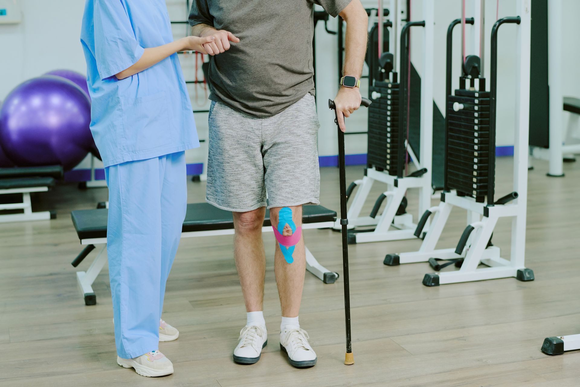 A physical therapist assists a man with a cane walking in a gym; man has knee tape.