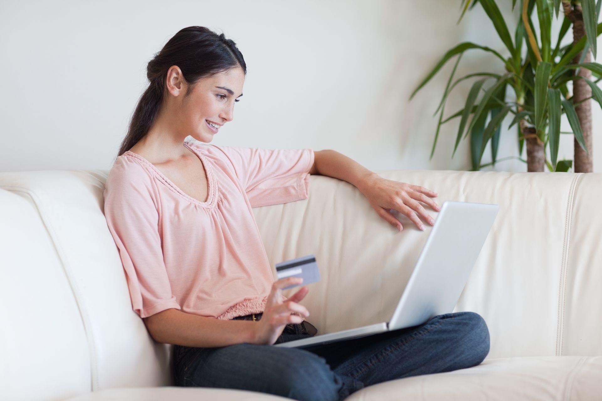 Woman on a white couch, smiling while entering credit card details on a laptop. Houseplant visible.