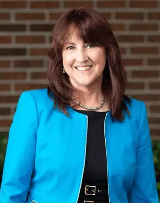 Woman with brown hair, smiling, wearing a blue jacket and necklace, in front of a brick wall.