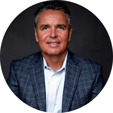 Woman with brown hair, smiling, wearing a blue jacket and necklace, in front of a brick wall.