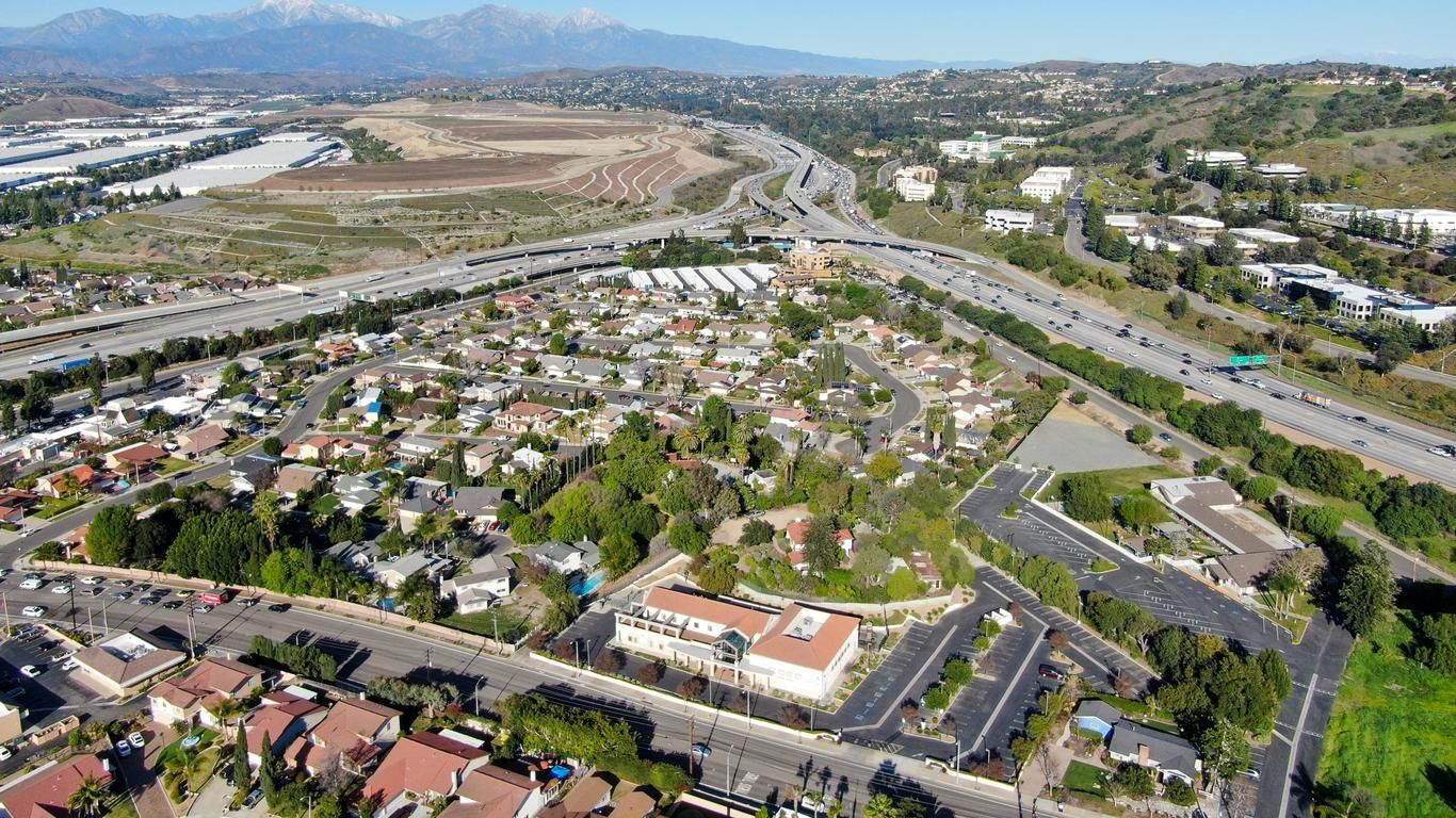 Aerial view of a suburban neighborhood beside a highway and a wide dry riverbed, with mountains in the distance.