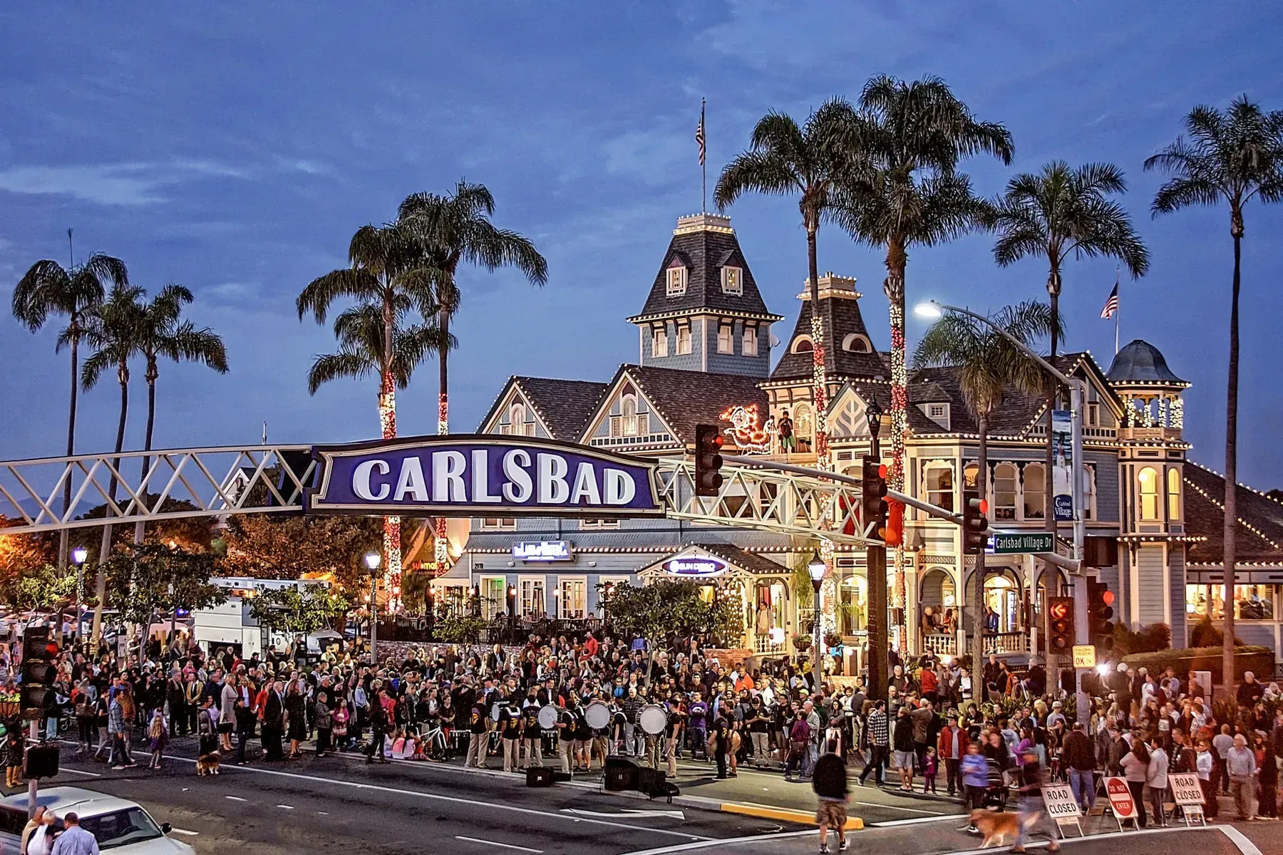 Crowded Carlsbad festival entrance at dusk, with illuminated archway, palm trees, and historic-style buildings