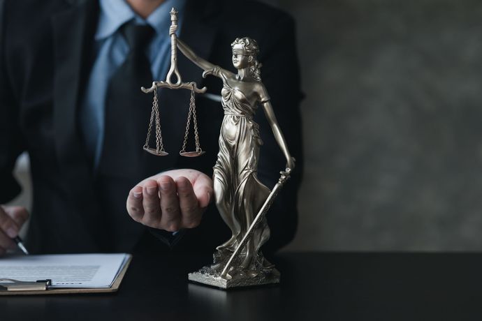 Lawyer in suit with blindfolded Lady Justice statue holding scales and sword. Clipboard on desk.