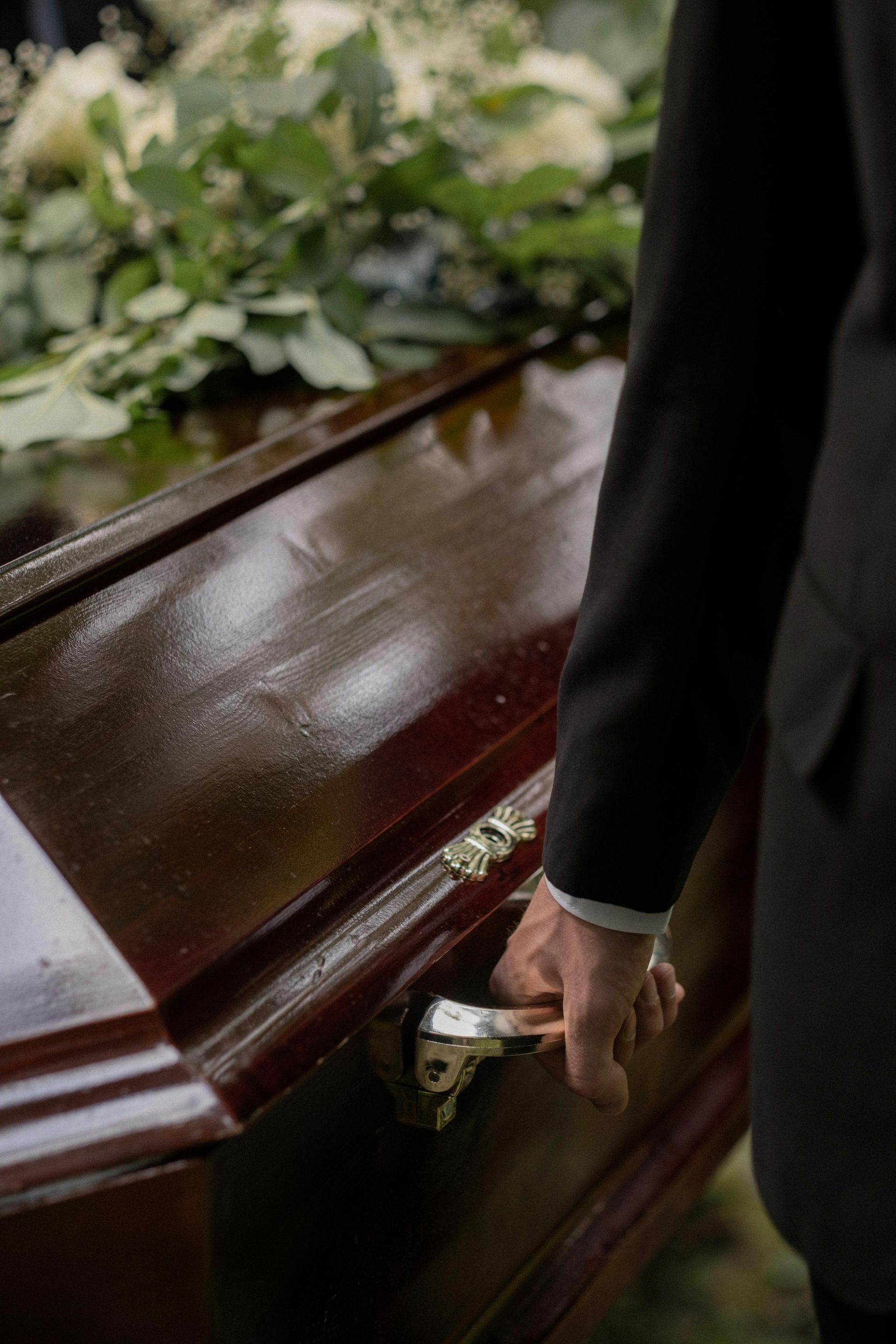 Person in suit holding coffin handle, white flowers in background.