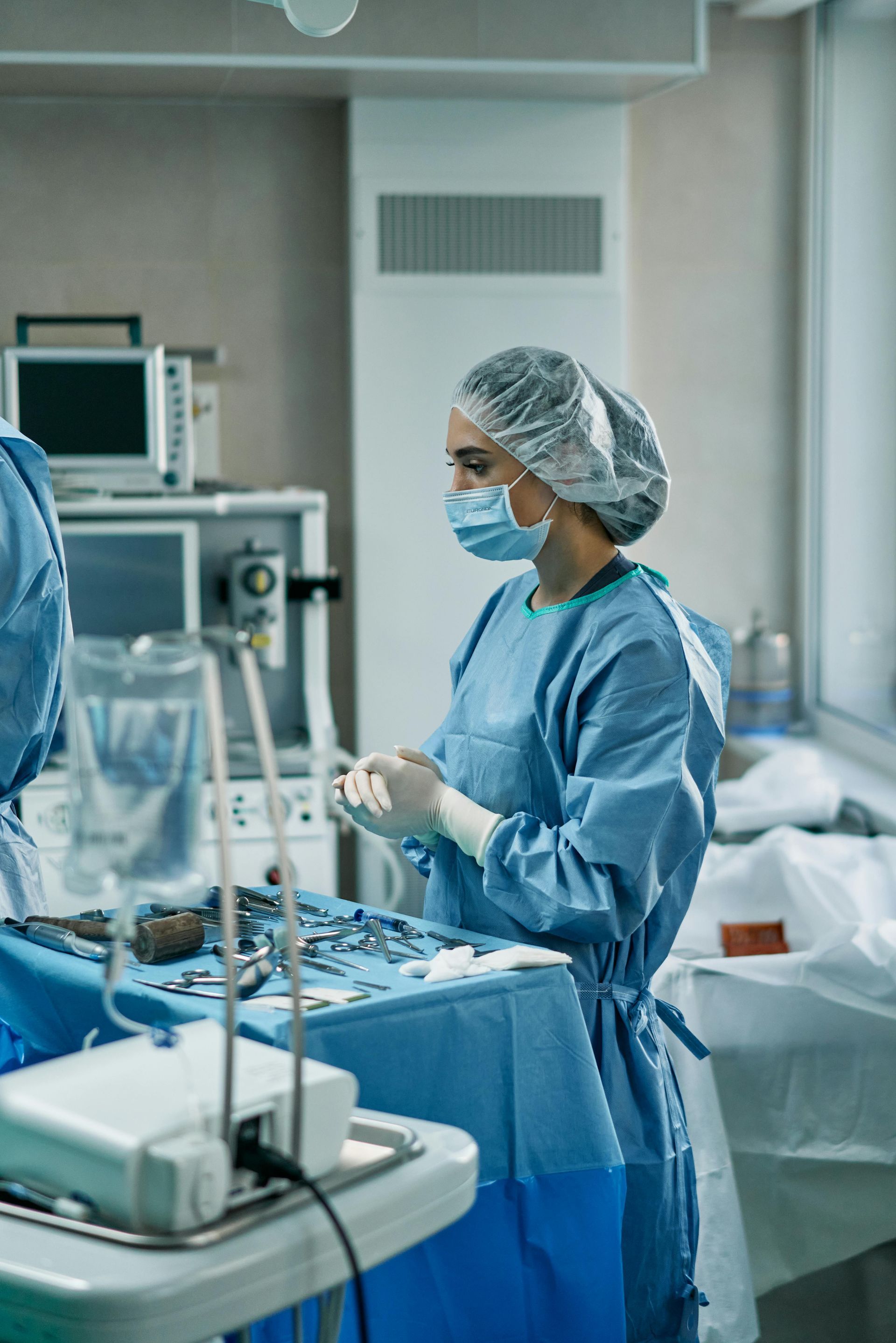 Surgeon in blue scrubs, mask, and cap prepares surgical instruments in an operating room.