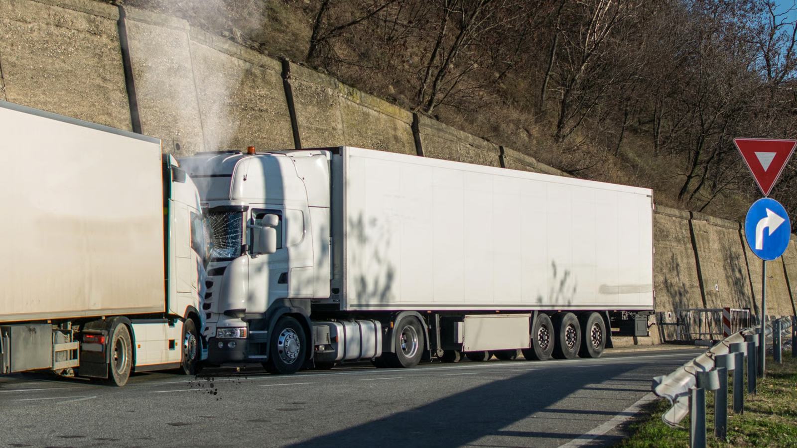 Two white semi-trucks parked on a roadside beside a concrete wall and traffic signs