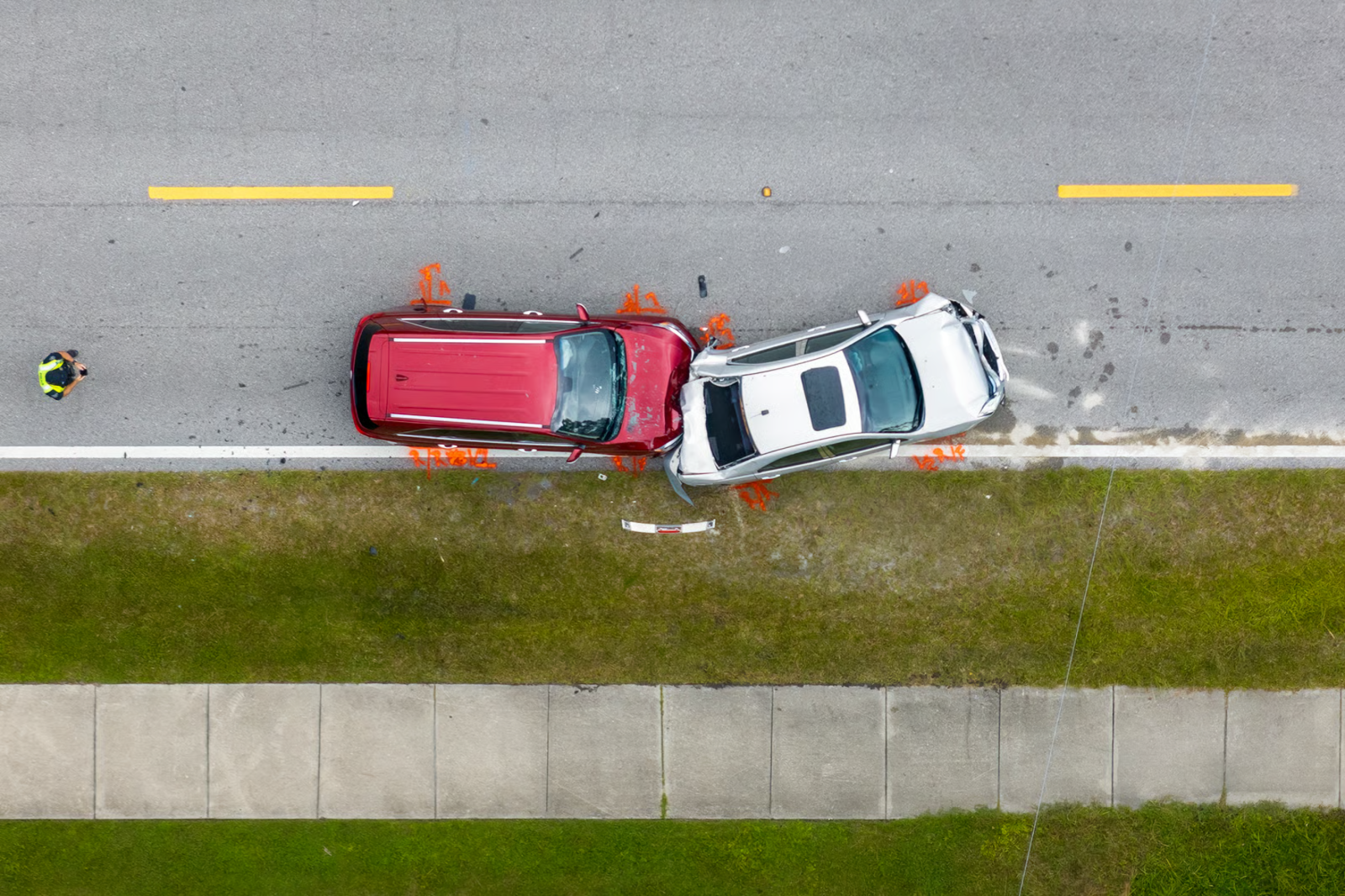 Overhead view of a red and a white car crashed on a roadside beside a sidewalk and grass