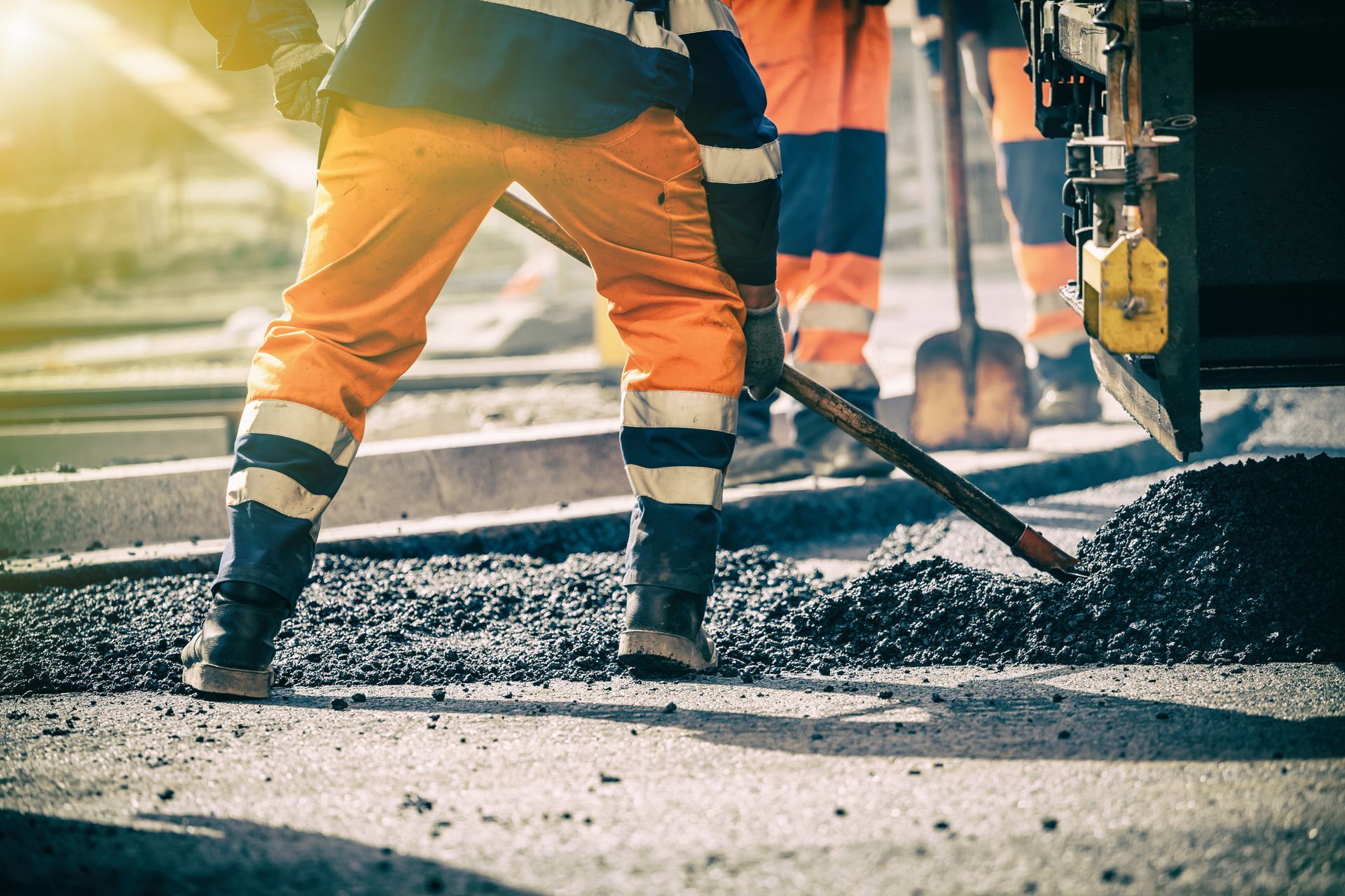 A construction worker shovels asphalt, wearing orange and blue reflective gear, by a paving machine.
