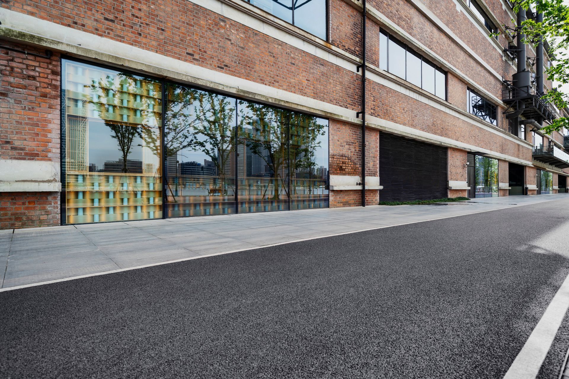 Exterior view of a brick building with large glass windows reflecting trees and a black asphalt road.