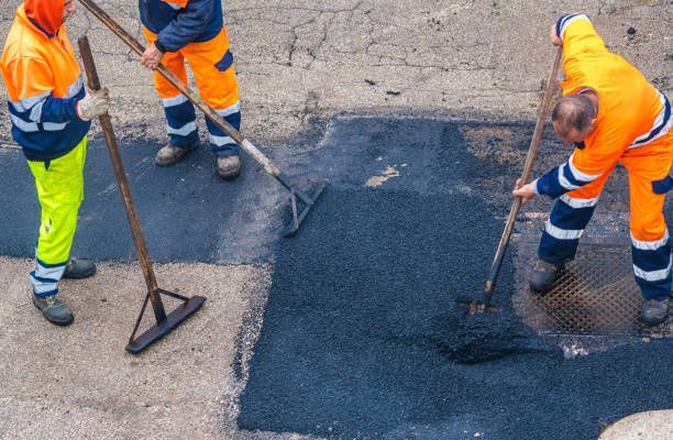 Three road workers in orange and blue coveralls, raking asphalt on a city street.