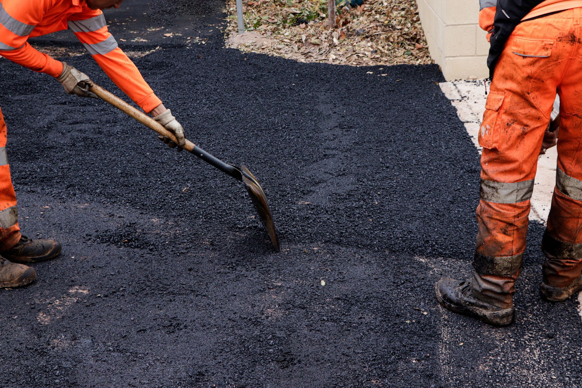 Two workers in orange coveralls spread asphalt with shovels.