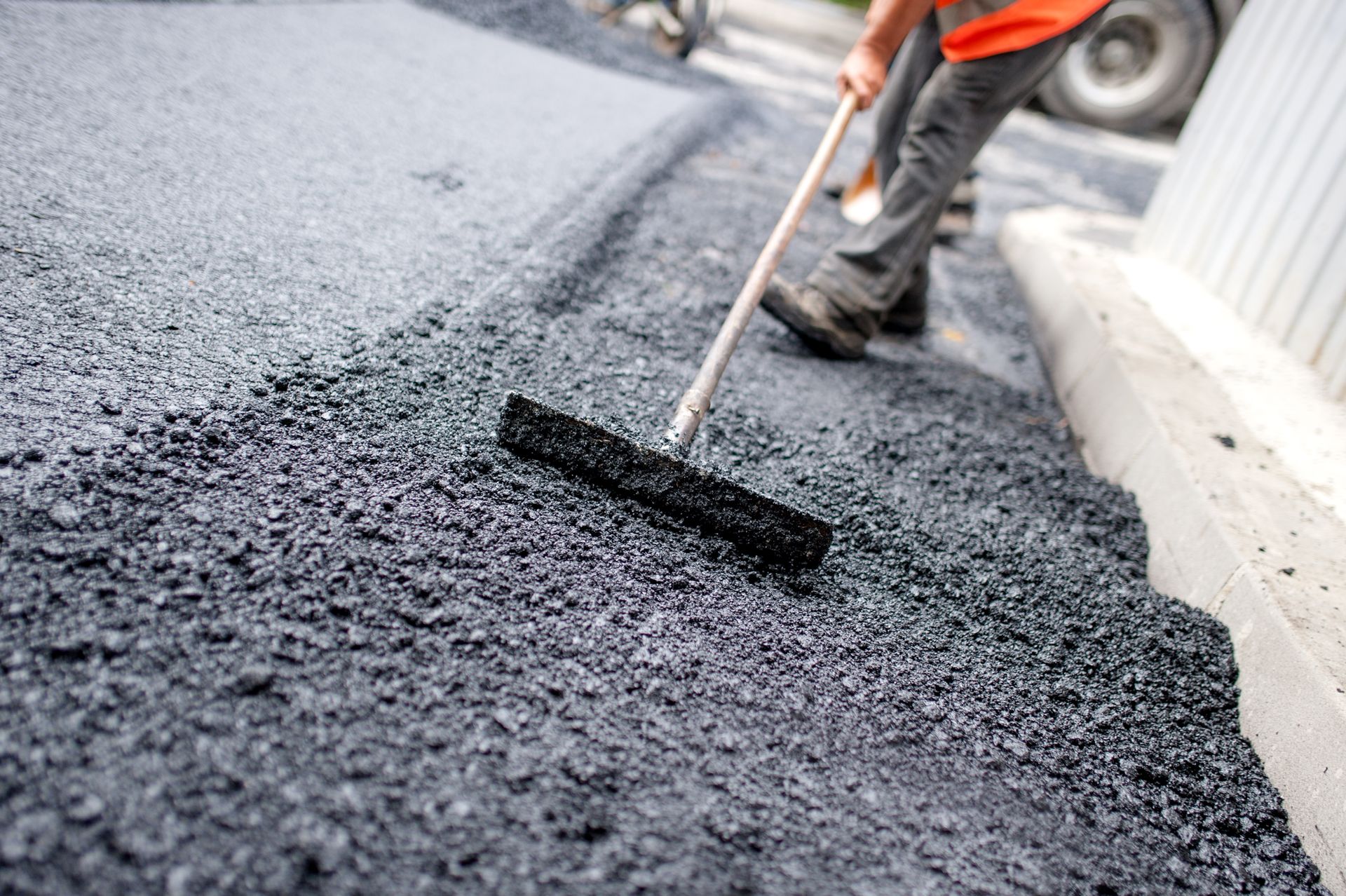 Person in orange vest using a broom to spread fresh asphalt on a road.