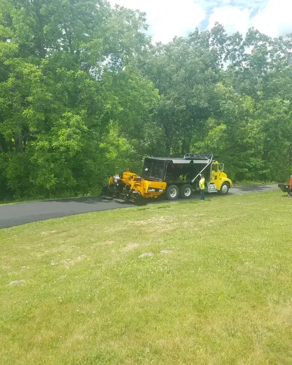 Asphalt paving in progress: dump truck feeding asphalt paver on a road, trees in background, sunny day.