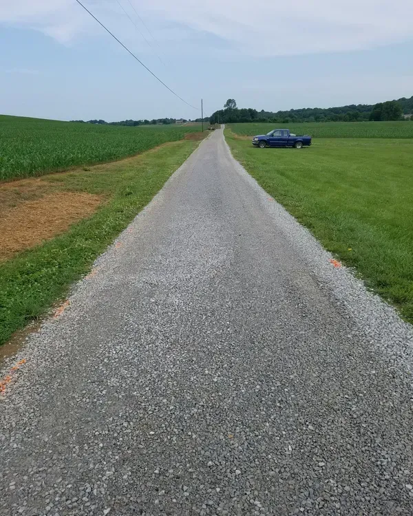 A gravel road extends through fields of grass and crops, a blue car is parked on the right.