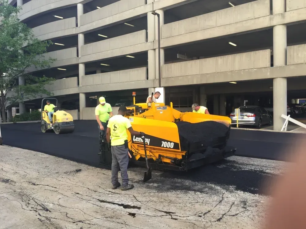 Asphalt paving crew at work near a parking garage. Yellow paver, workers in safety vests, roller.