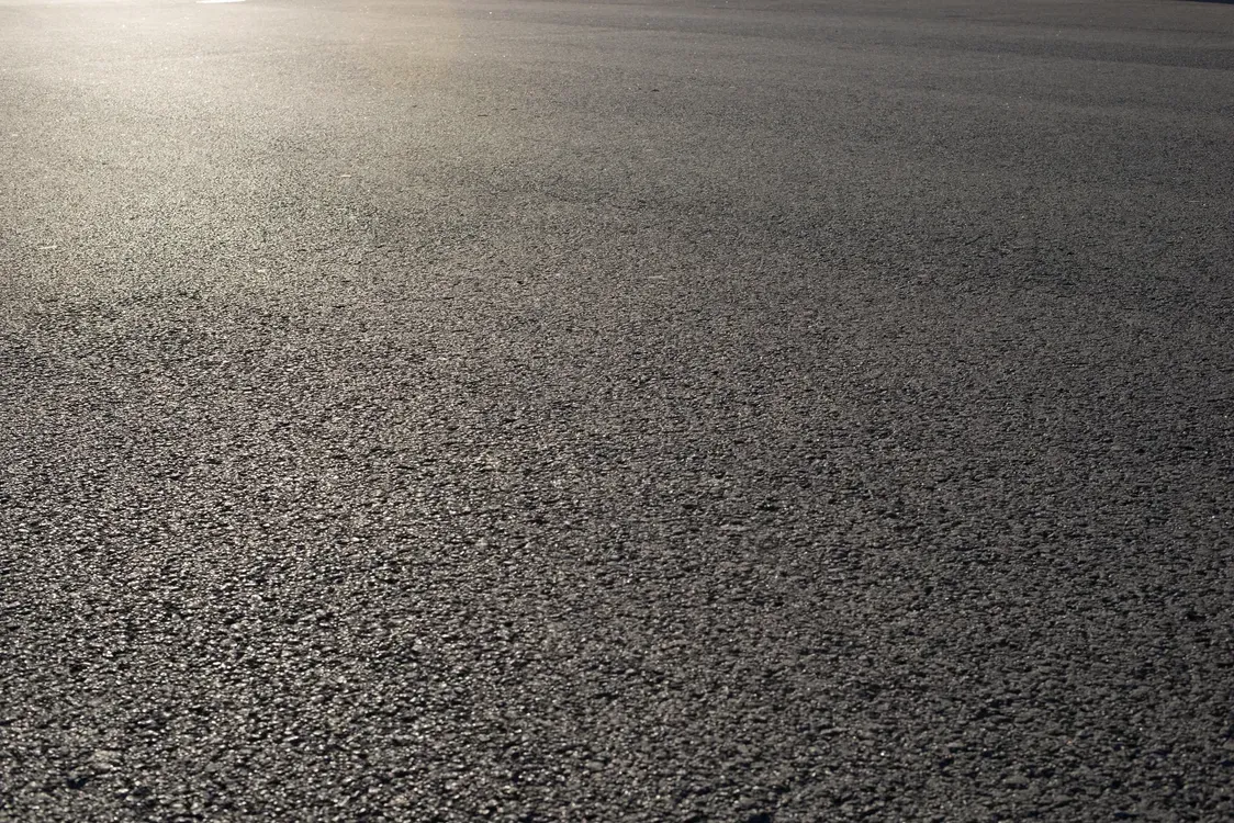 Close-up view of asphalt pavement, showing a rough, textured surface with sunlight reflecting.