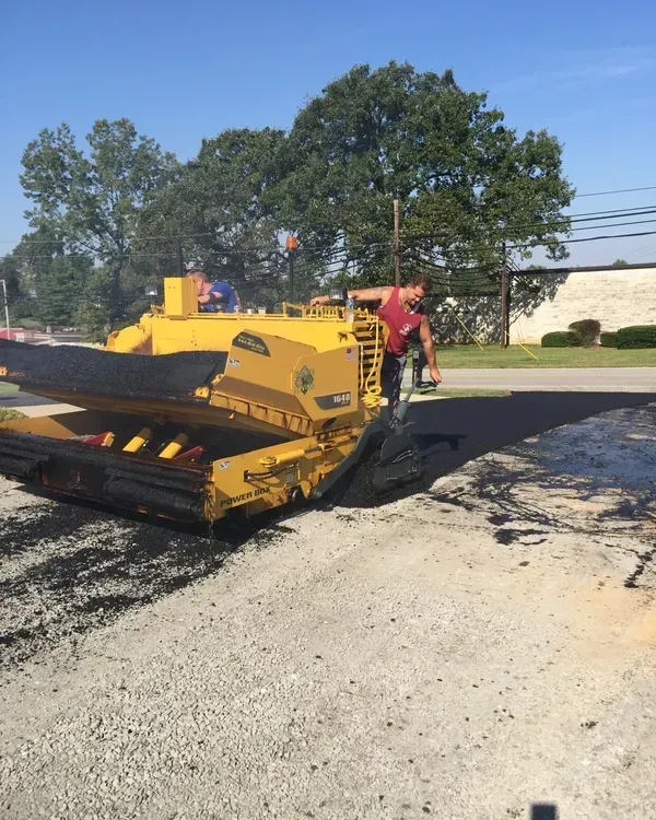 Yellow asphalt paving machine laying new pavement on a road. Man in red shirt works alongside.