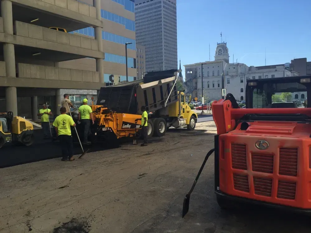 Road construction: Workers in safety vests paving a street with asphalt, next to a yellow dump truck and orange machinery.