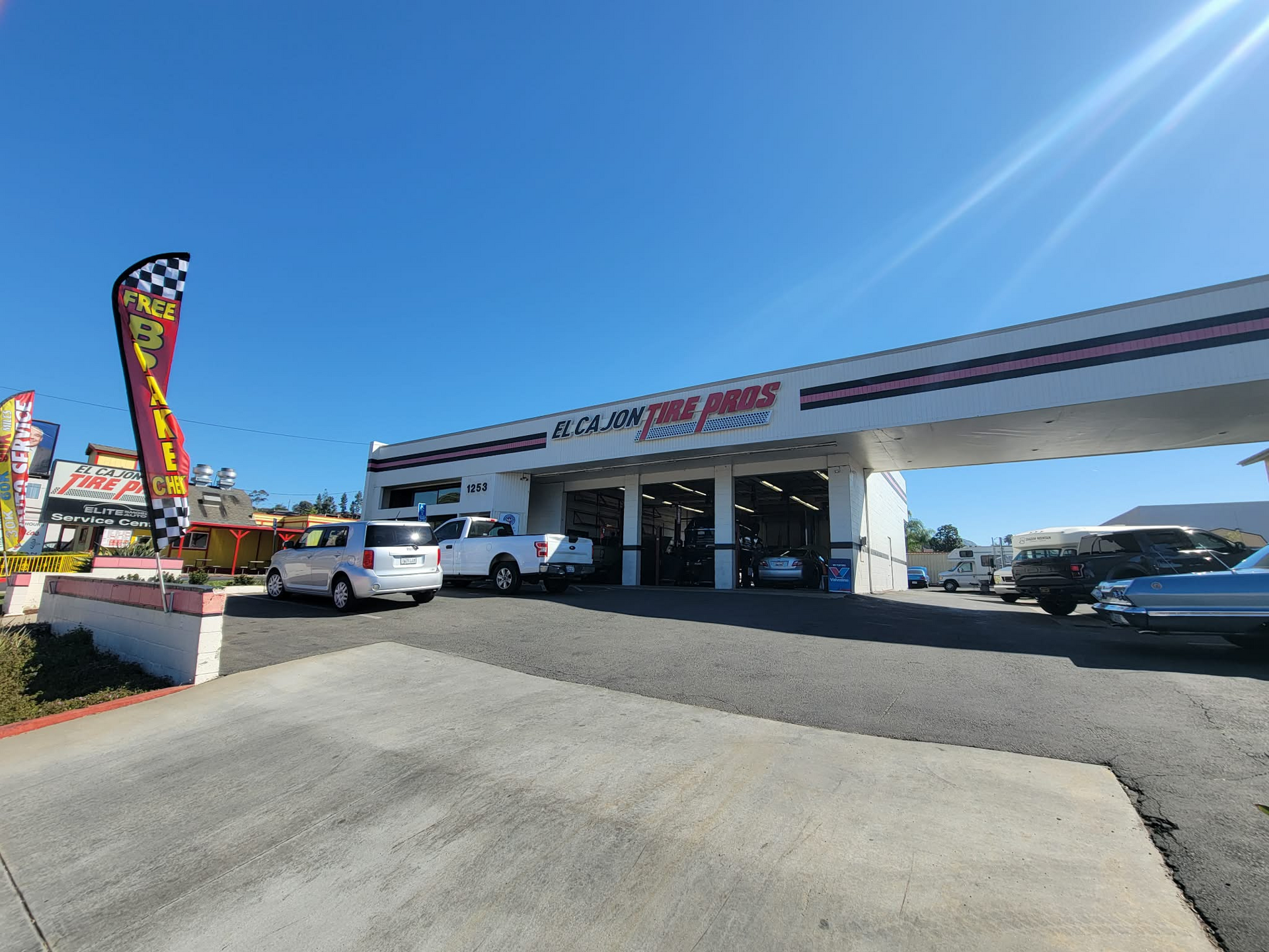 A wide-angle shot of a white tire shop building under a clear blue sky, with a tall, colorful vertical sign in the front. | El Cajon Tire