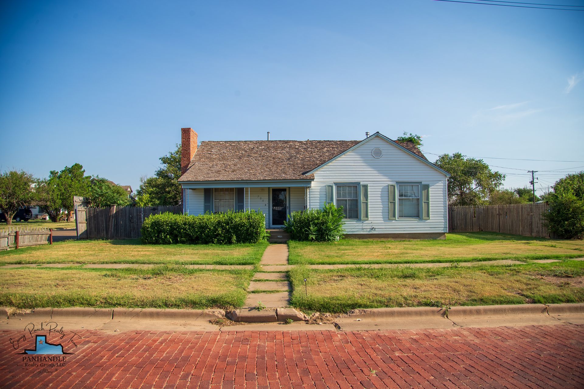 Small white house with light green shutters, brown roof, brick sidewalk, and a clear blue sky.