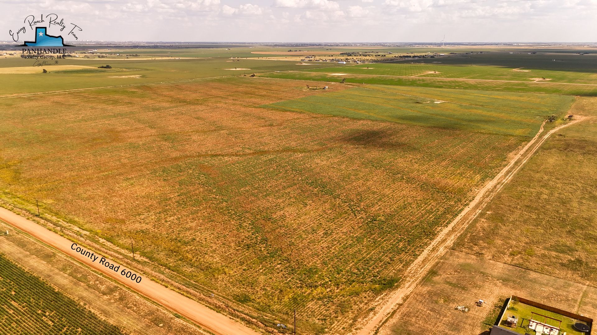 Vast open field with dirt roads, patches of green, and a distant building under a cloudy sky.