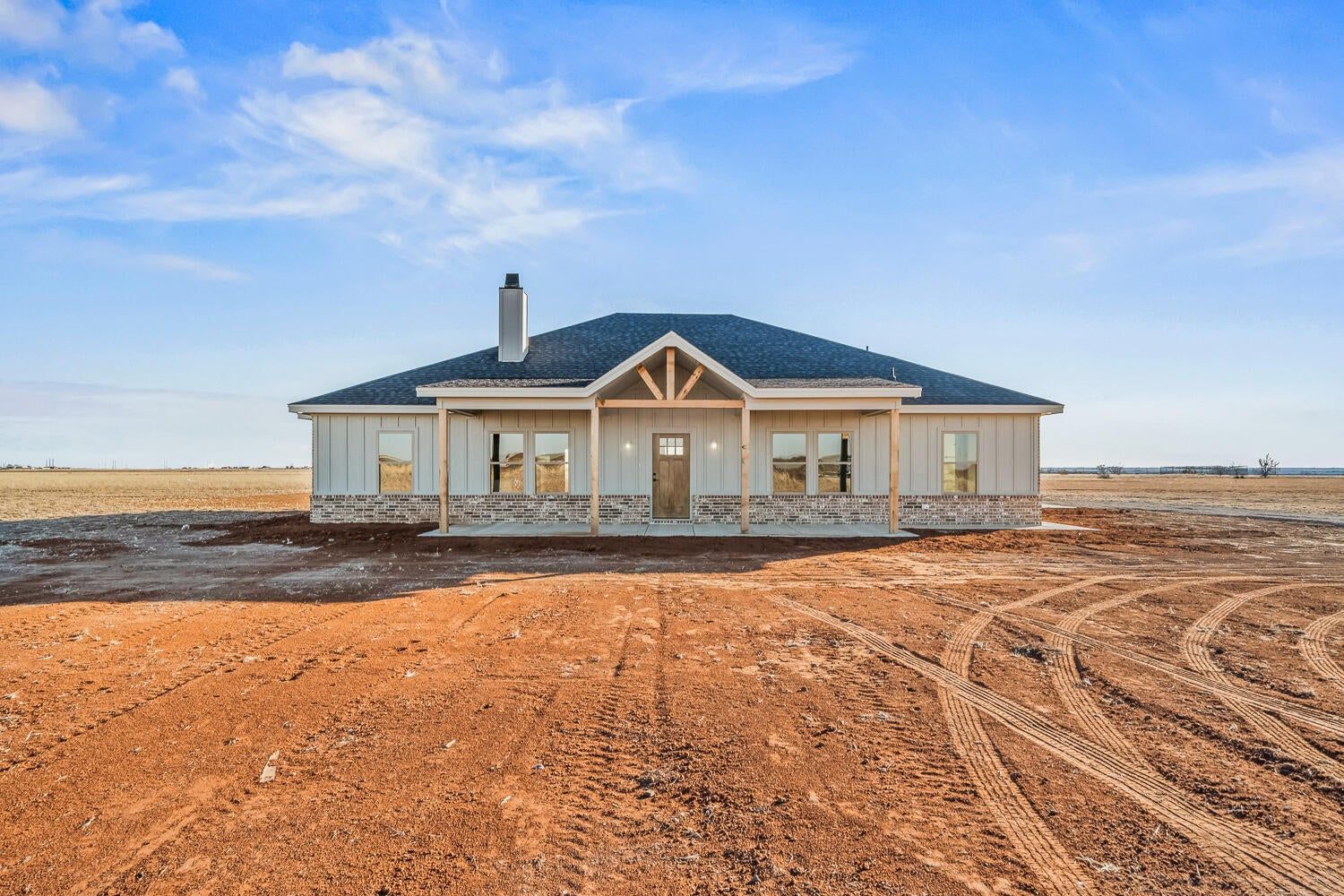 A ranch-style house with a black roof sits on red dirt under a blue sky.