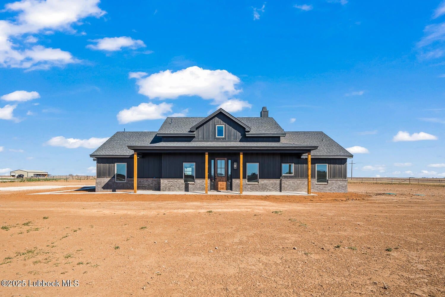 Dark-colored ranch house with porch and pillars on a large, empty lot under a bright blue sky.