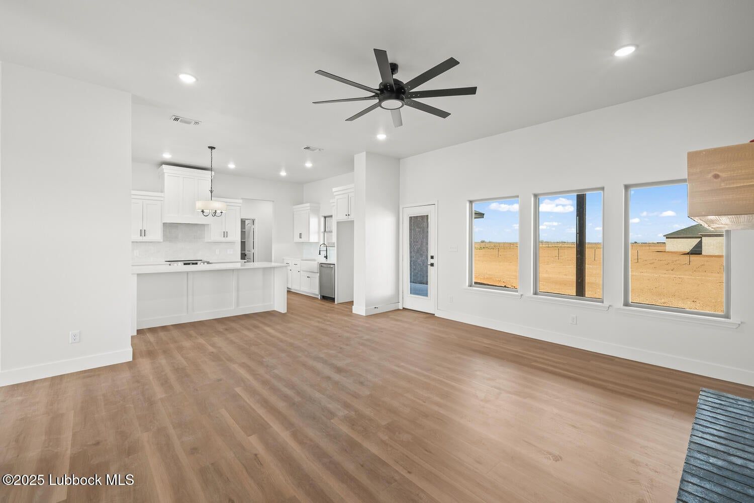 Spacious, bright living room with wood floors, white walls, and open-concept kitchen. Windows show a desert landscape.