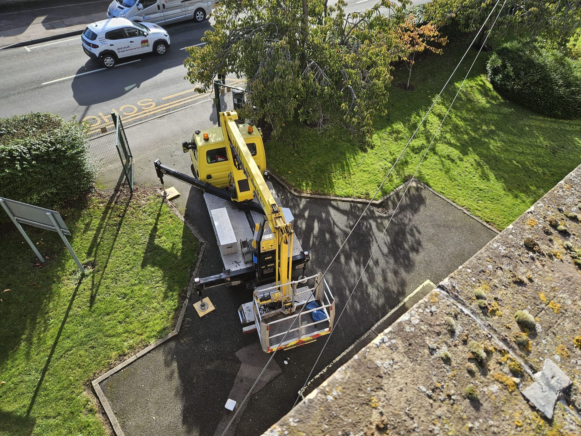 Yellow crane on a driveway near a road. A white car drives by. Green grass and a building's edge are also visible.