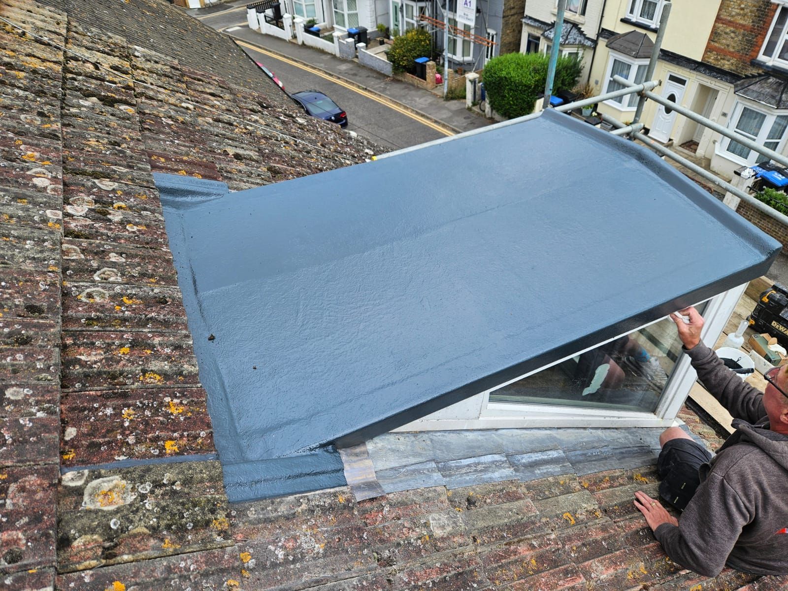 A man is sitting on top of a roof holding a piece of roofing material.