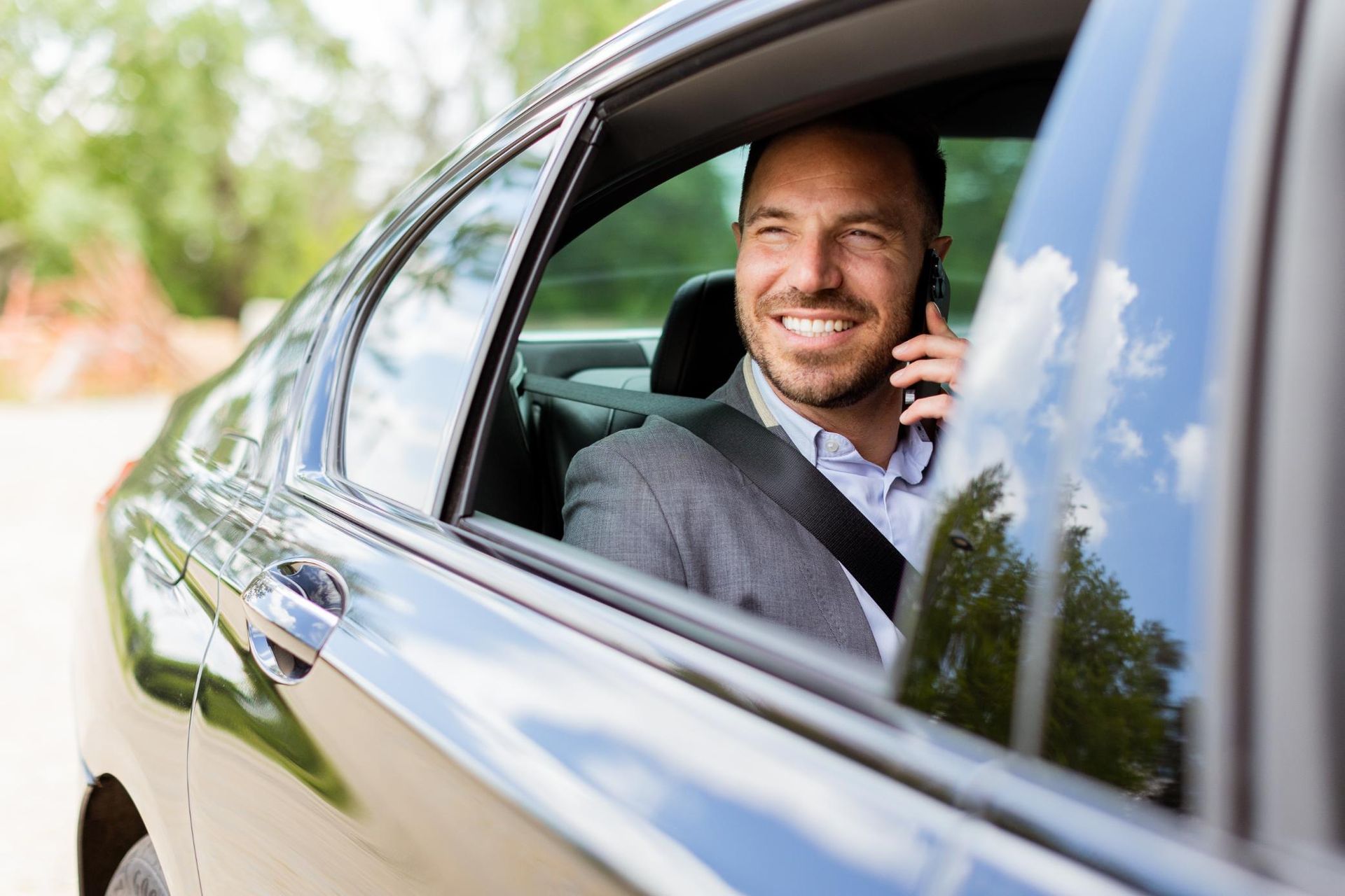 Man smiling in a car window while talking on a phone, with trees reflected in the glossy black door